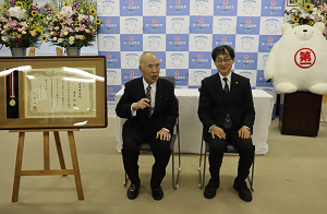 上=大分大学名誉博士称号と第一交通産業のマスコット、タクマンに囲まれる形で会話をする黒土始・第一交通産業創業者会長(左)と北野正剛・大分大学学長(右)